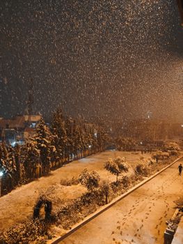 A serene winter night with snow falling on an empty city street, illuminated by streetlights.