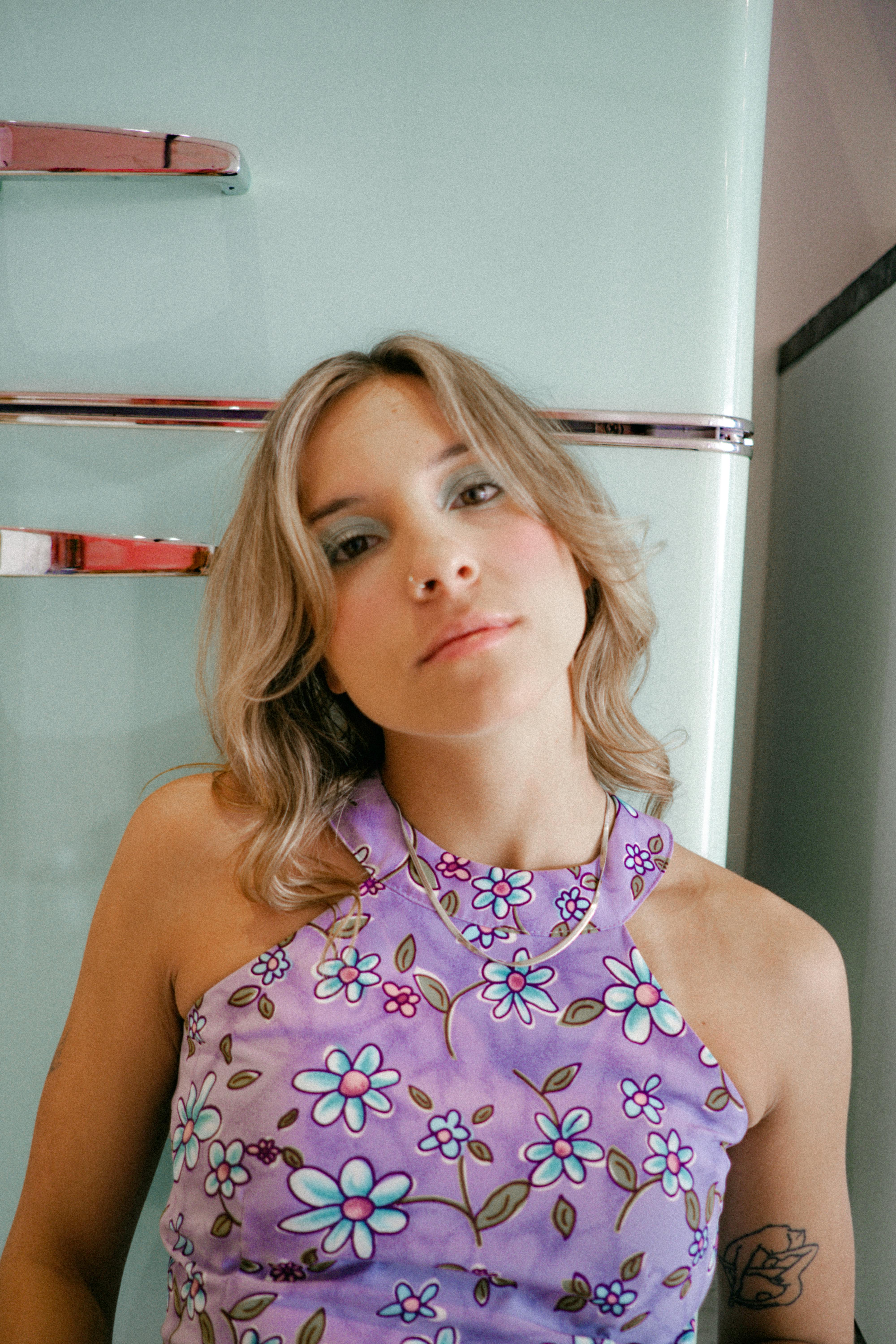 Young woman in a floral dress posing indoors with vintage style fridge. Soft light and pastel tones.