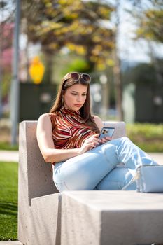 Woman sitting on a concrete bench in a sunny park, using her smartphone with headphones on.
