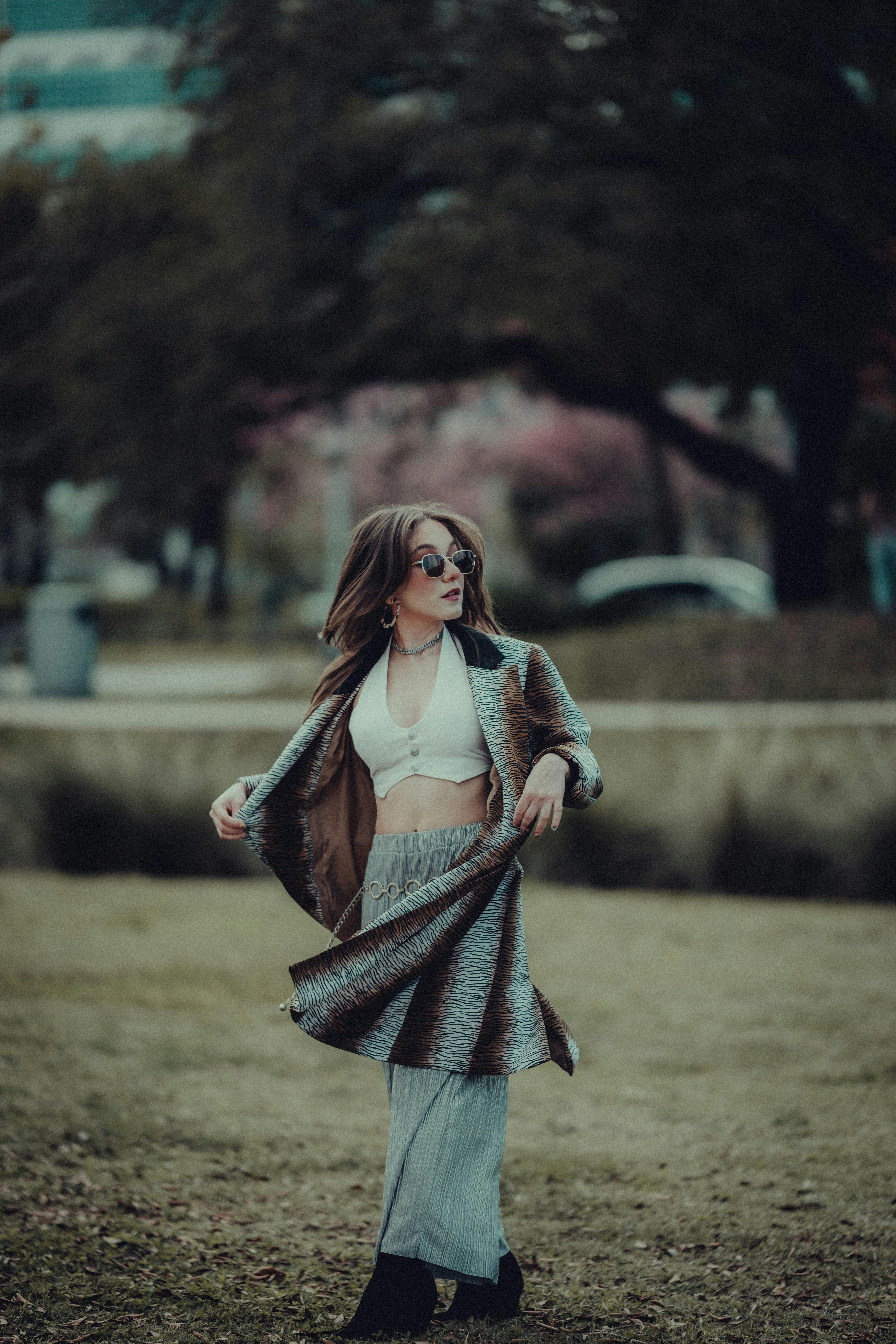 Fashionable woman in stylish outfit posing in a park, wearing sunglasses and a coat.