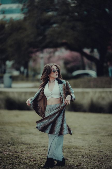 Fashionable woman in stylish outfit posing in a park, wearing sunglasses and a coat.