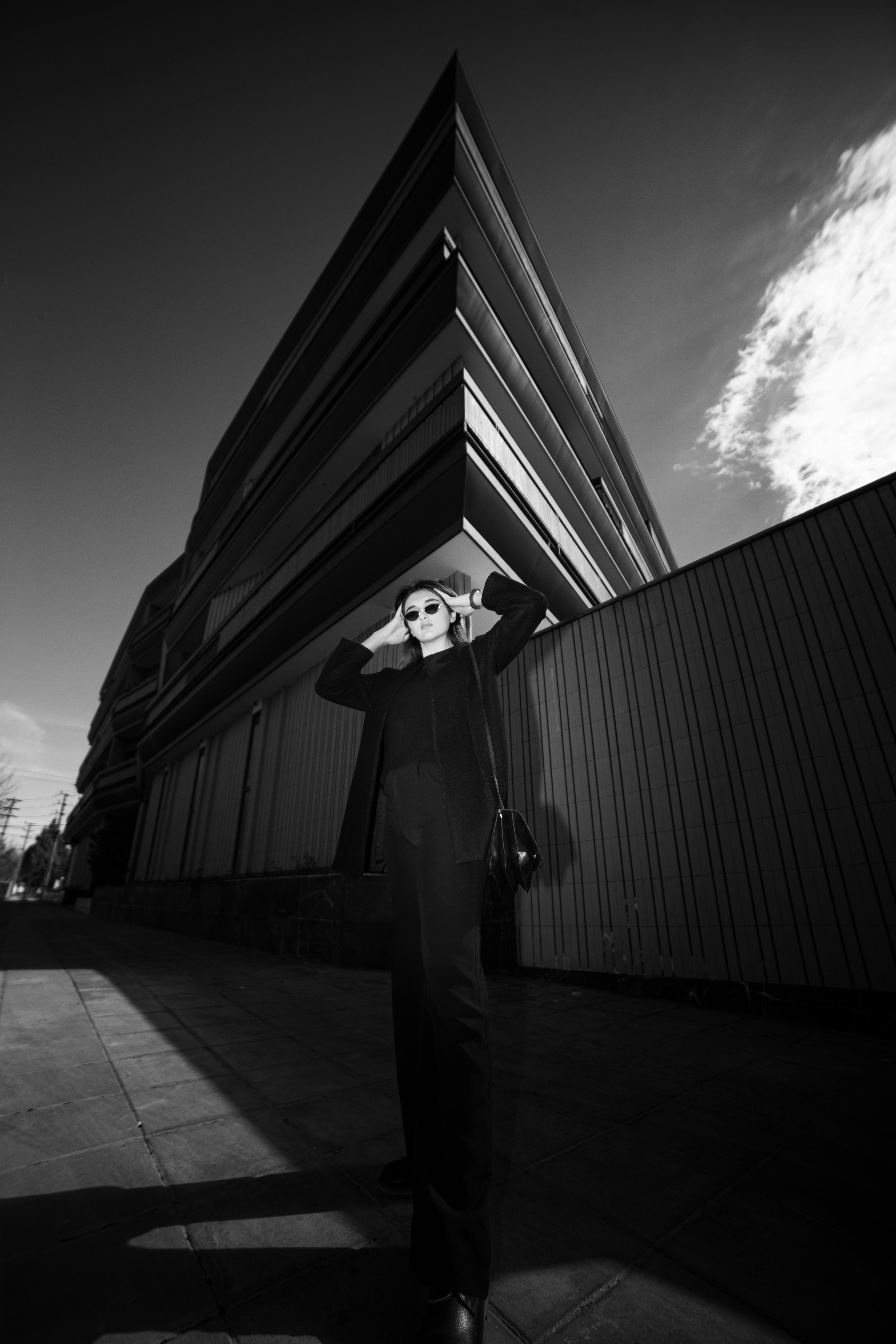 Stylish woman in black attire poses confidently near modern urban architecture in a high-contrast black and white composition.