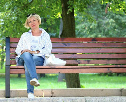 Thinking Woman in White Jacket and White Scoop Neck Shirt Blue Denim Jeans Sitting on Brown Wooden Bench Beside Green Trees during Daytime