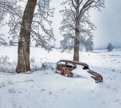 A vintage car covered in snow sits abandoned in a snowy field with frost-covered trees in Minnesota.