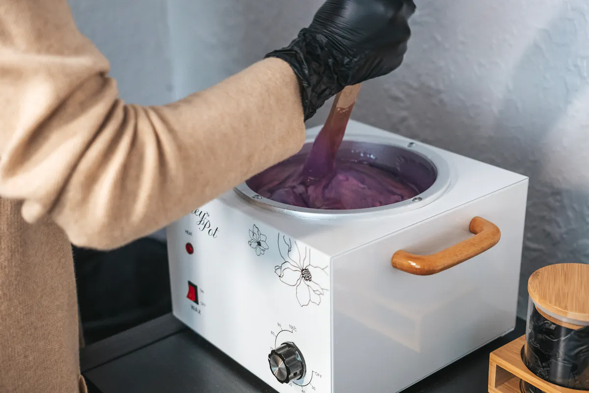 Close-up of a gloved hand stirring purple wax in a professional wax warmer, preparing for waxing services like leg waxing and bikini wax for hair removal.