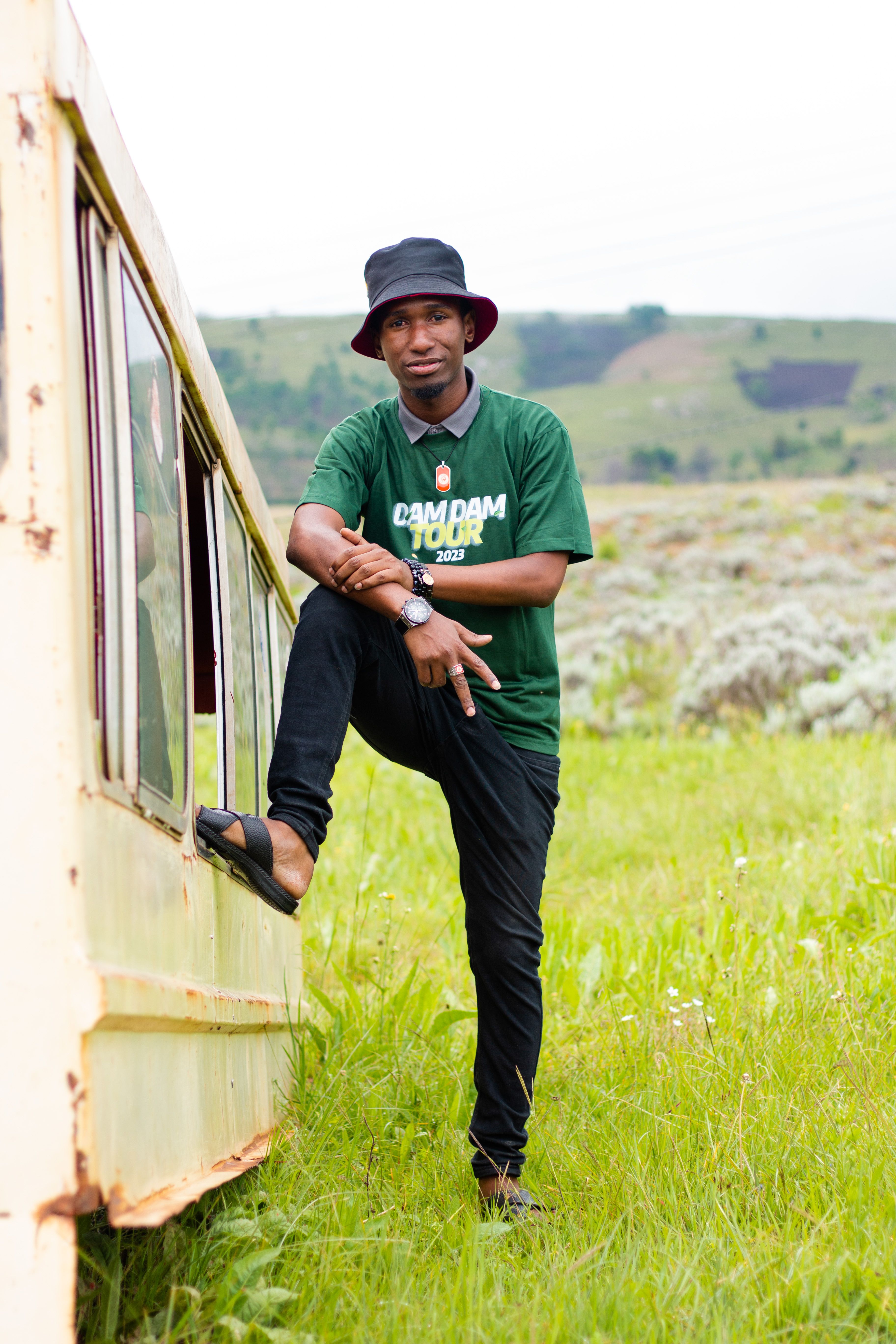 A young man in casual clothing leans against an abandoned train car in a lush countryside setting.