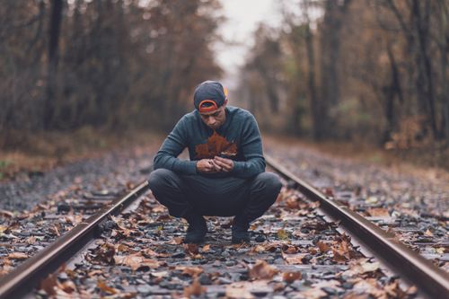 Free A man squats on train tracks surrounded by autumn leaves, wearing a sweater and baseball cap. Stock Photo