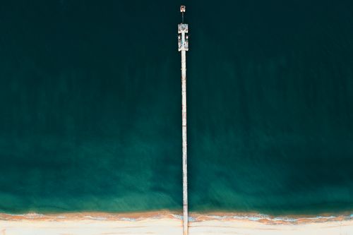 Free A long pier stretches from the sandy beach into the vast ocean from a bird's-eye view. Stock Photo