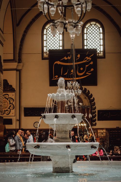 Free A beautiful stone fountain in a bustling mosque interior with people admiring the architecture. Stock Photo