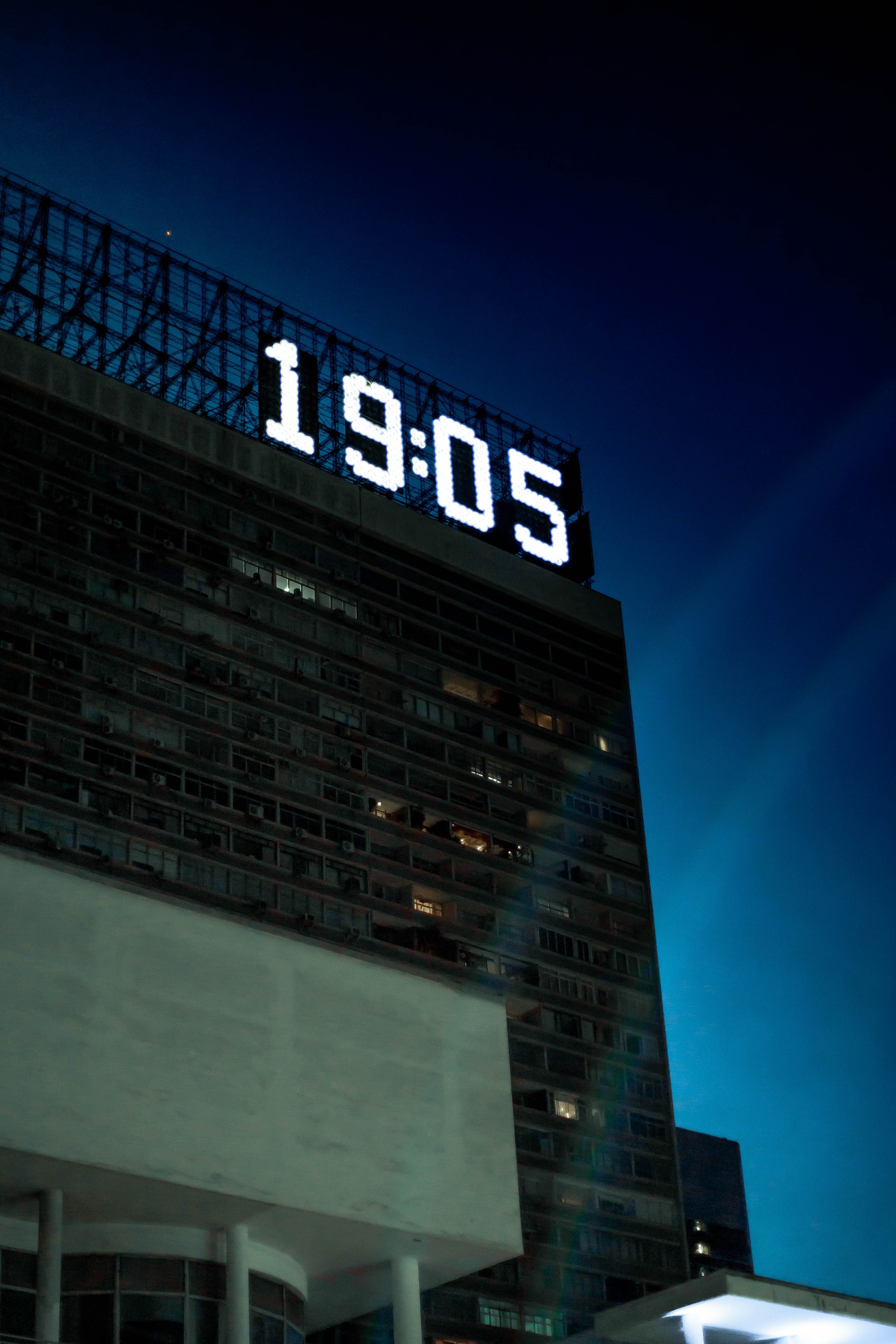 Modern office building with large illuminated digital clock displaying 19:05 against a twilight sky.