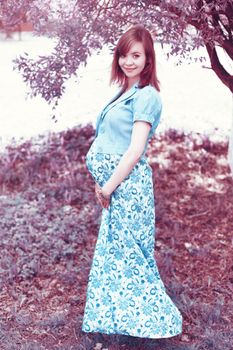A happy pregnant woman in a floral dress smiles under a tree, capturing a joyful moment.