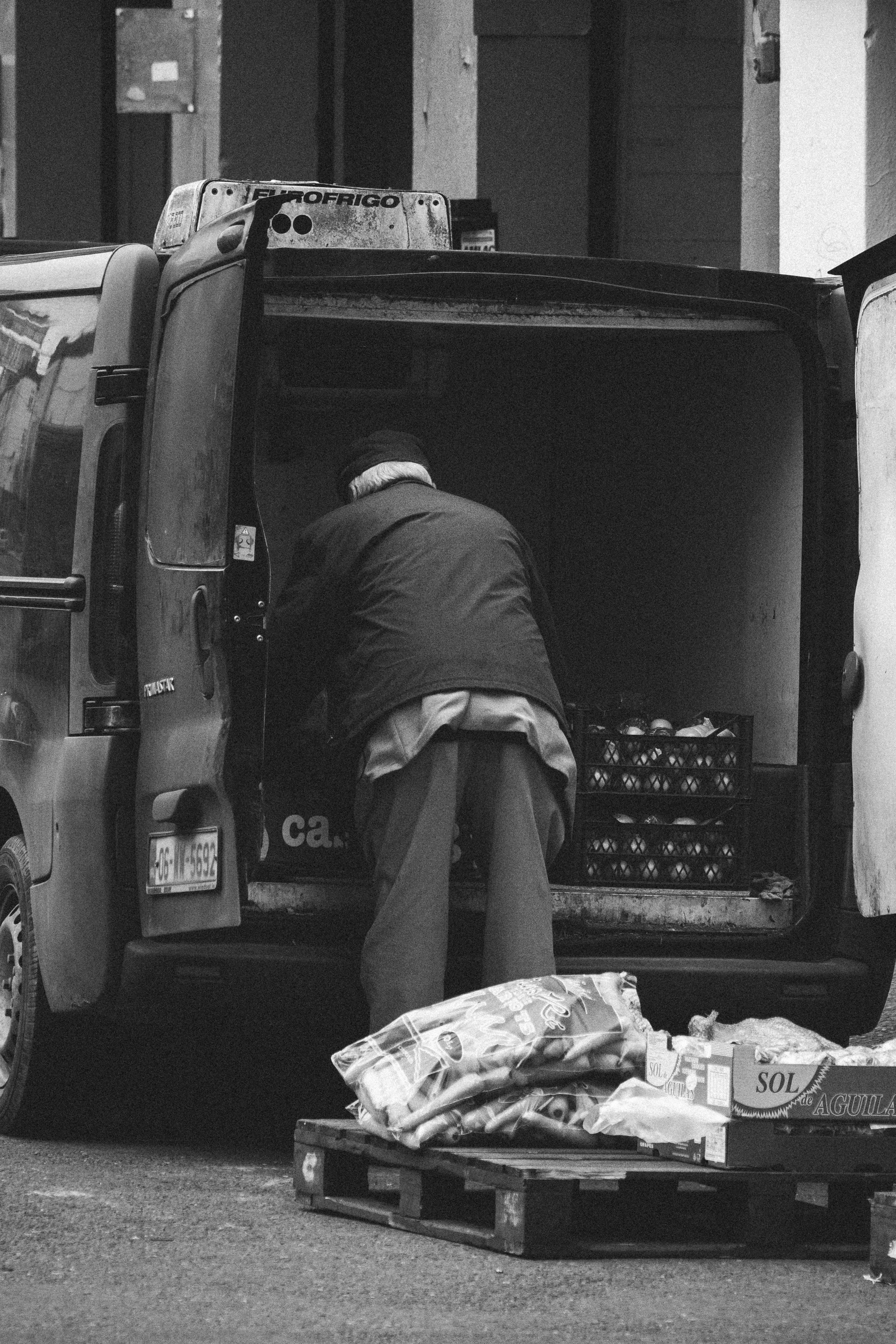 Black and white image of an elderly man unloading pallets of crates from a van.