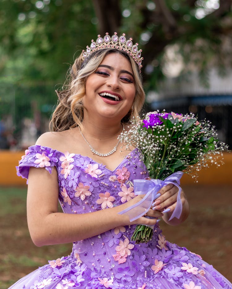 Photo Of A Young Woman In A Purple, Floral Dress And A Tiara, Holding A Bouquet And Smiling 