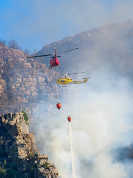 Two helicopters extinguishing a wildfire in a mountainous area, showcasing aerial firefighting efforts.
