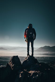 A lone hiker stands on a rocky peak, overlooking breathtaking mountains at sunrise in Afghanistan.