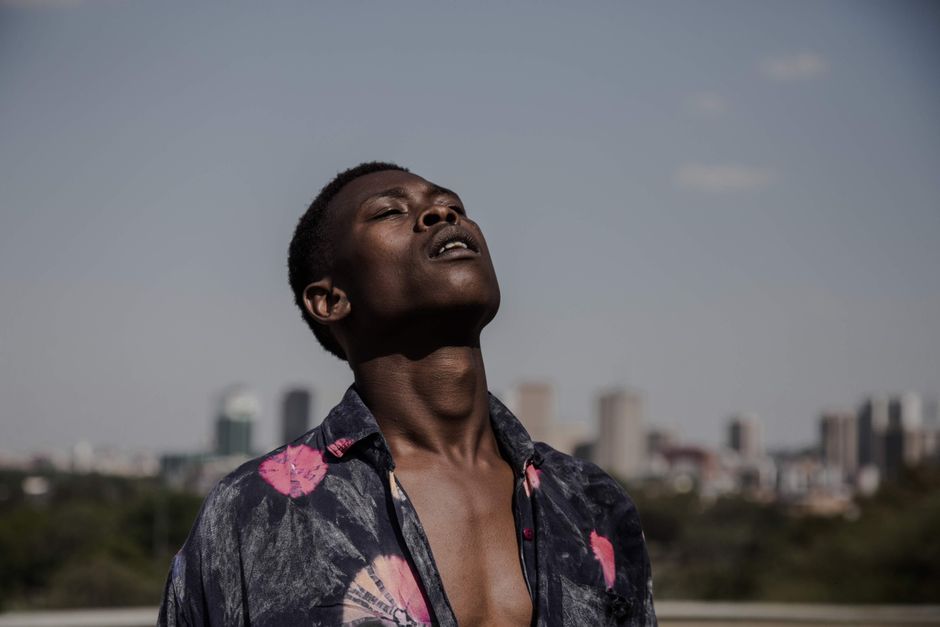 A young man in a floral shirt basks in daylight with a cityscape of Pretoria, South Africa, in the background.
