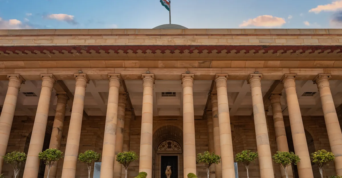 A government building in New Delhi with classical columns and the Indian national flag against a bright sky.