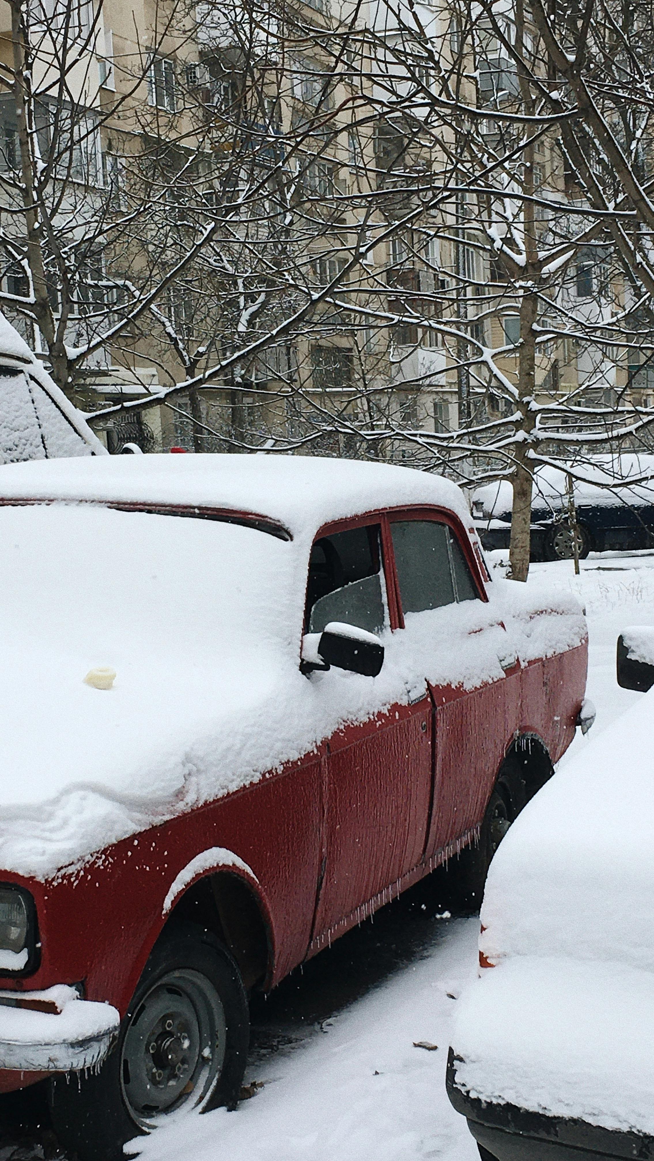 Red vintage car covered in snow, parked beside a snow-laden urban street in Odesa, Ukraine during winter.