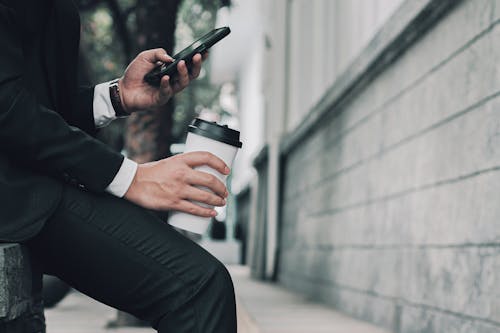 Close-up of a Man in a Suit Sitting on a Bench and Holding a Cup of Coffee and Using Phone 