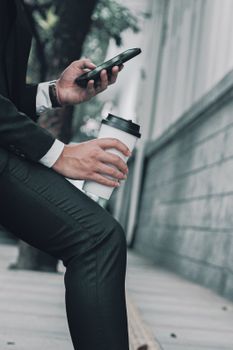 A man in a suit holding a coffee cup and smartphone while sitting outside.