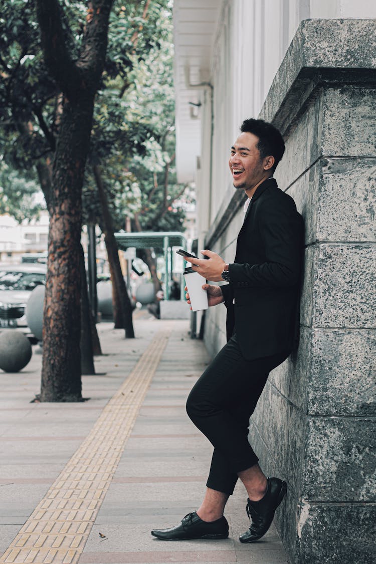 Laughing Man In Black Suit And Oxford Shoes Holding Coffee And A Smartphone Standing On The Sidewalk