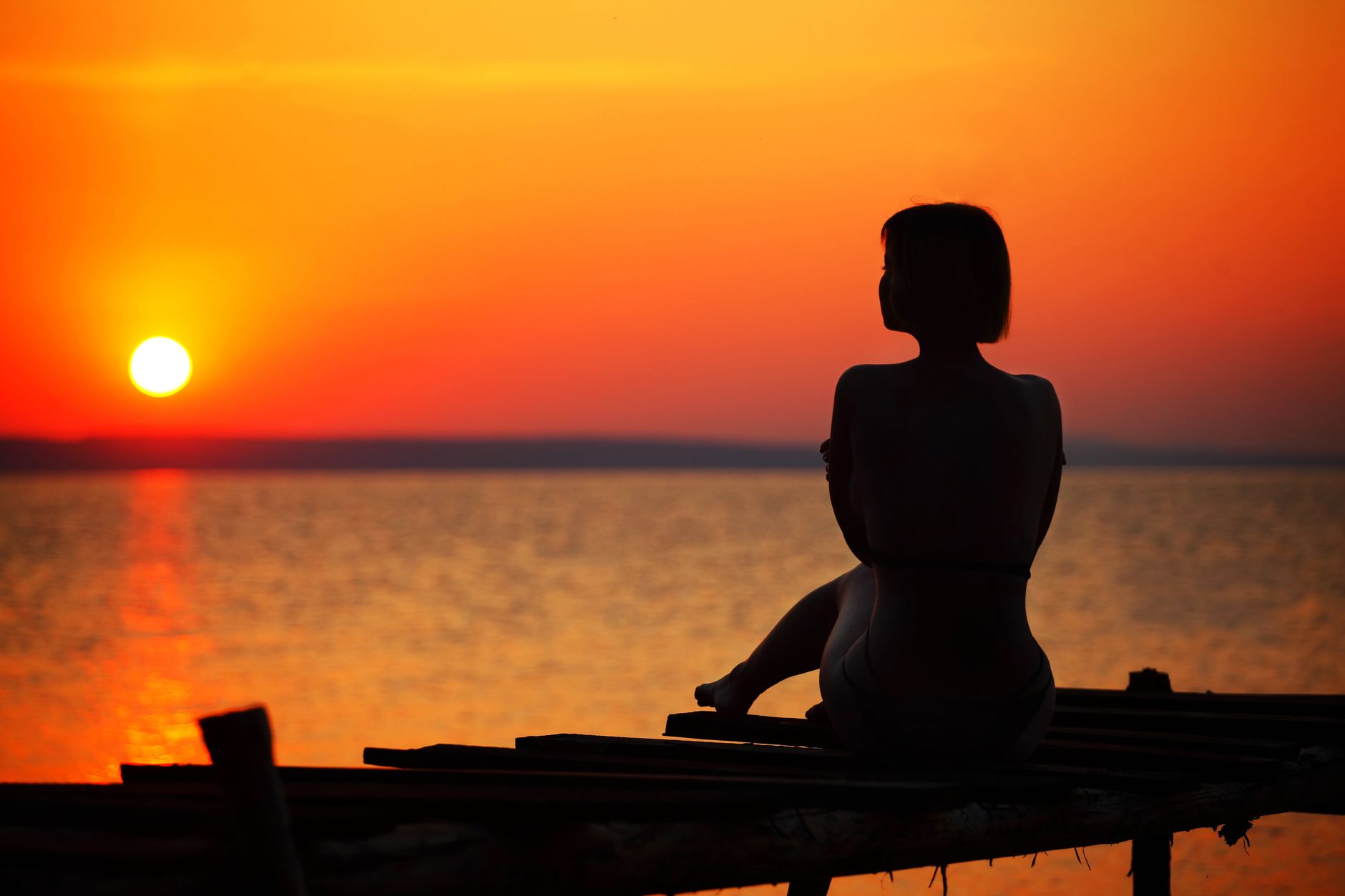 Serene silhouette of a woman sitting on a pier, watching the ocean at sunset.