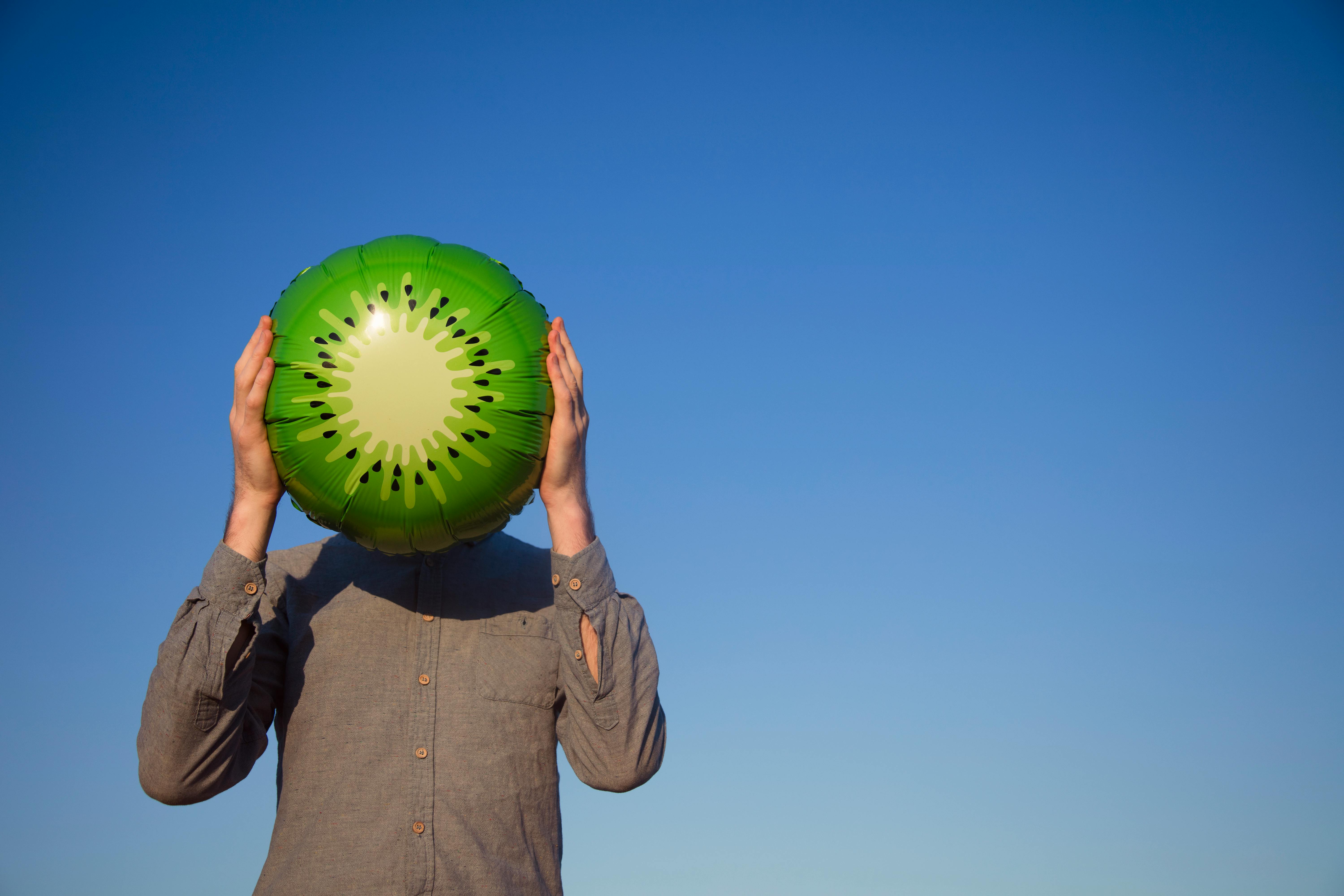 A man humorously holds a kiwi balloon in front of his head against a blue sky.