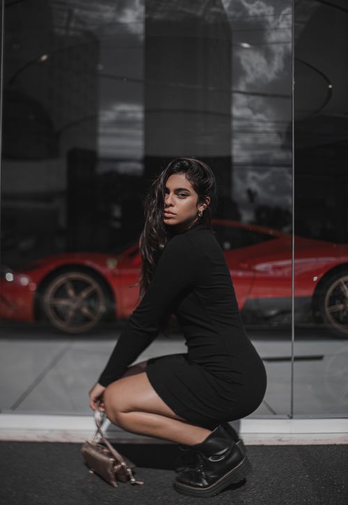 Free Elegant young woman in a black dress poses at a car showroom with a red sports car. Stock Photo