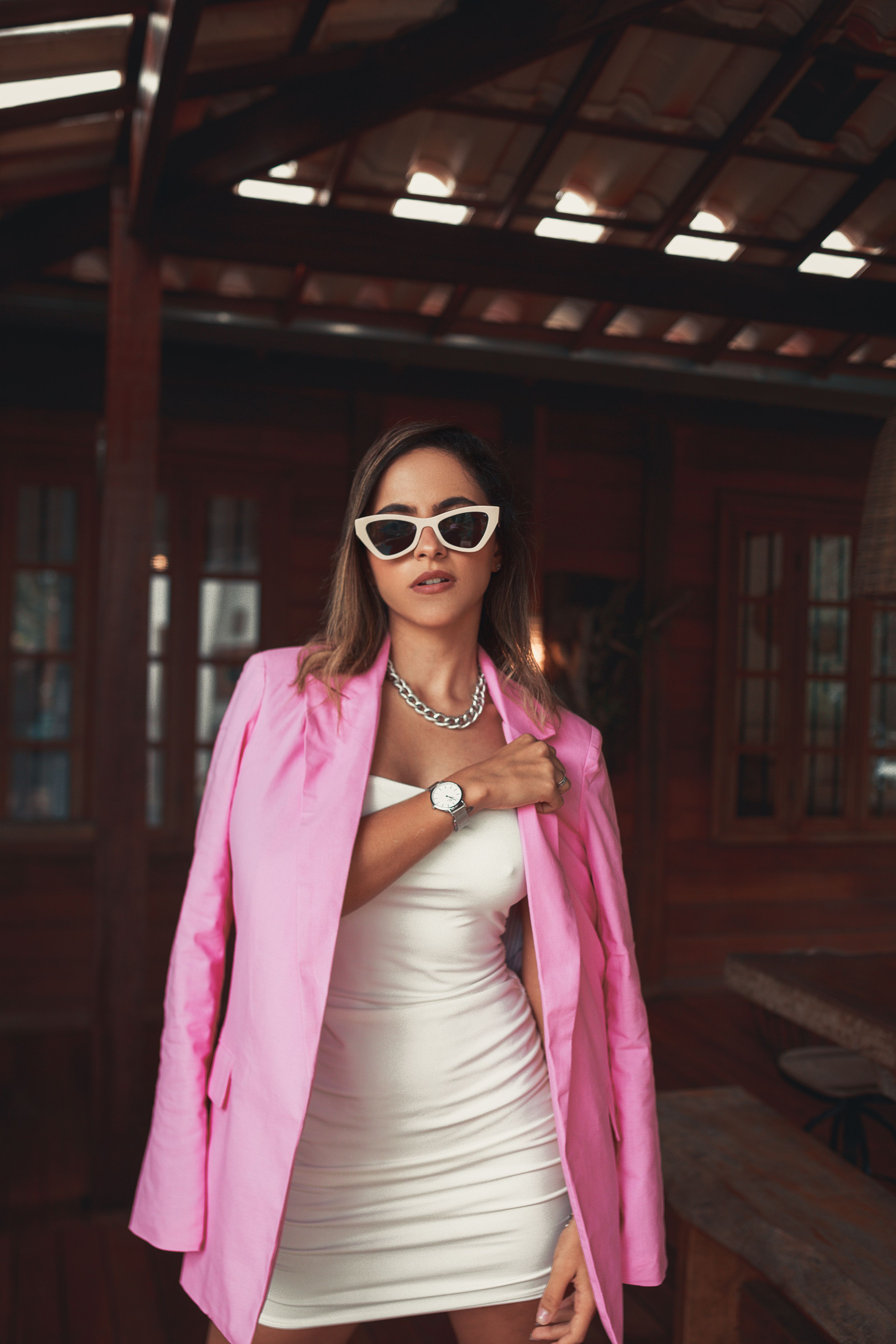 Mulher fashionista em um blazer rosa e vestido branco com óculos de sol, em pé dentro de casa.