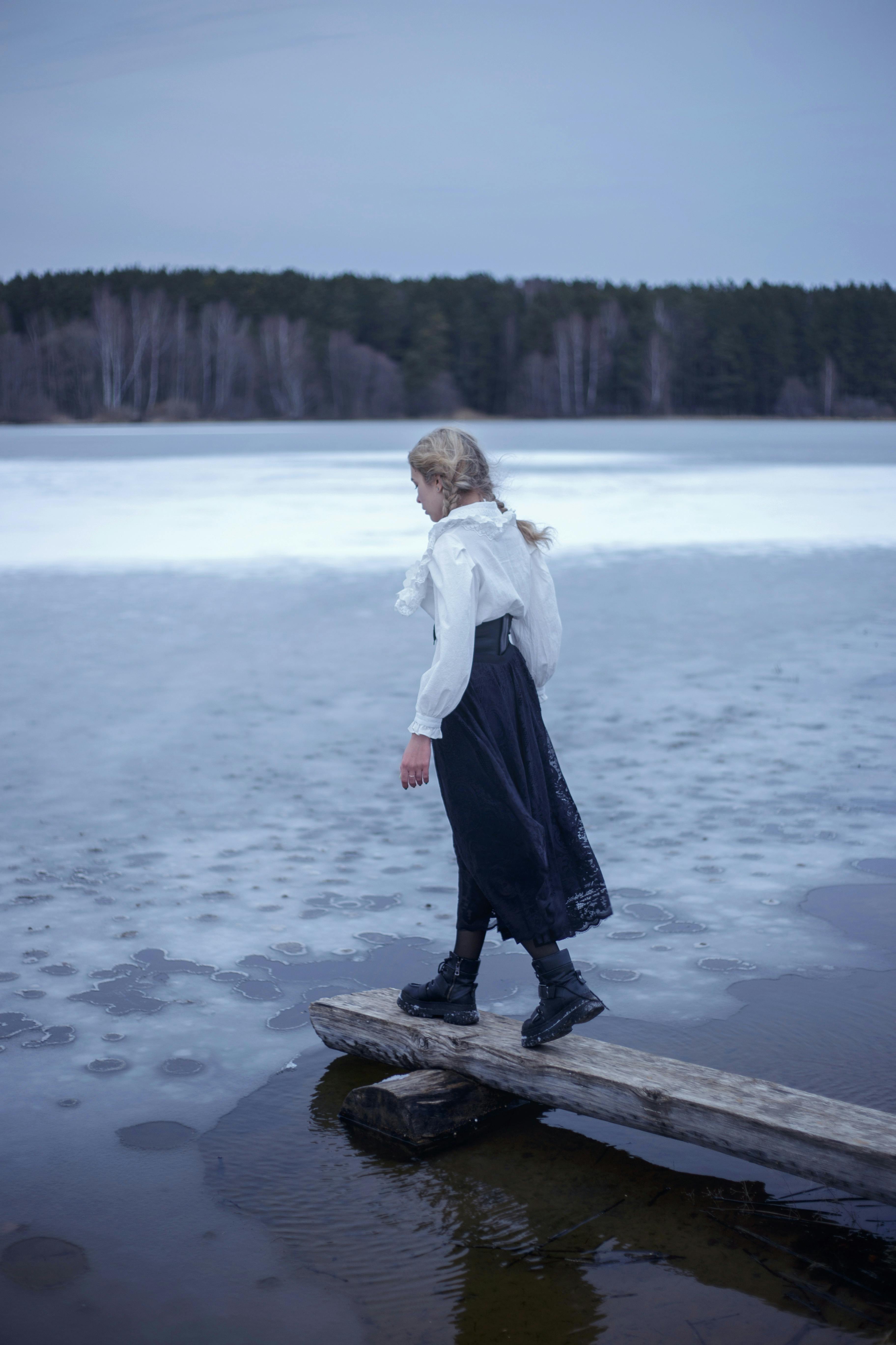 A woman carefully walking on a wooden board over a frozen lake in a serene winter scene.