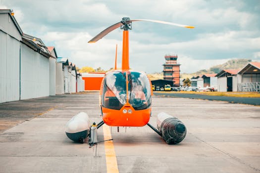 Bright orange helicopter on airstrip with airport towers and hangars in the background.