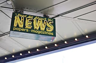 From below of illuminated signboard with news papers magazines inscriptions hanging on metal ceiling on street