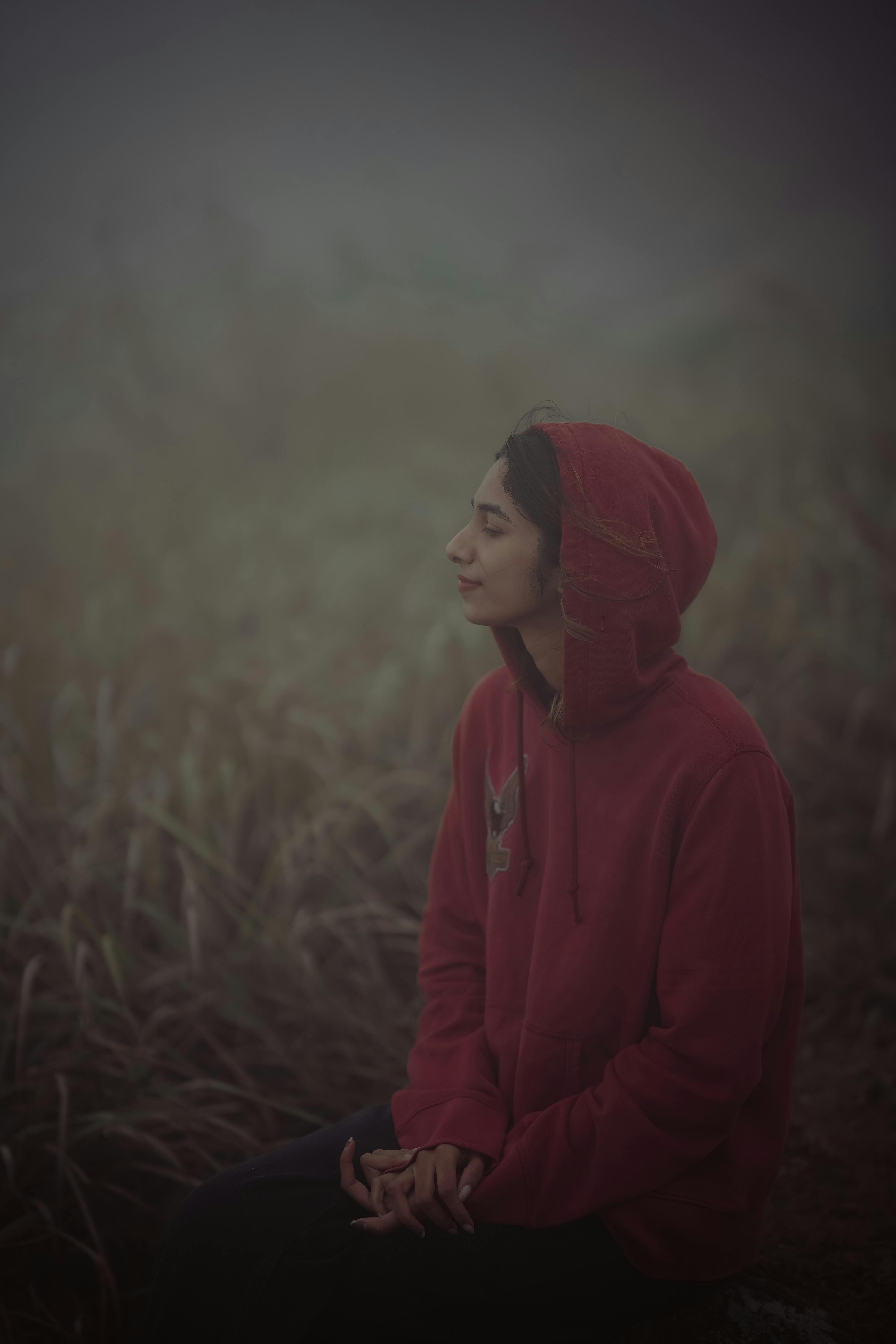 A woman sits in a red hoodie, meditating in foggy, grassy landscape.