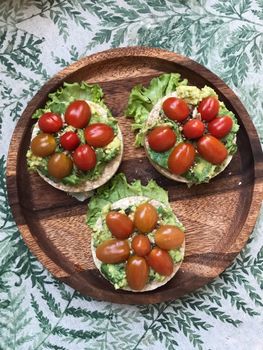 Top view of rice cakes topped with avocado, cherry tomatoes, and lettuce on a wooden plate.