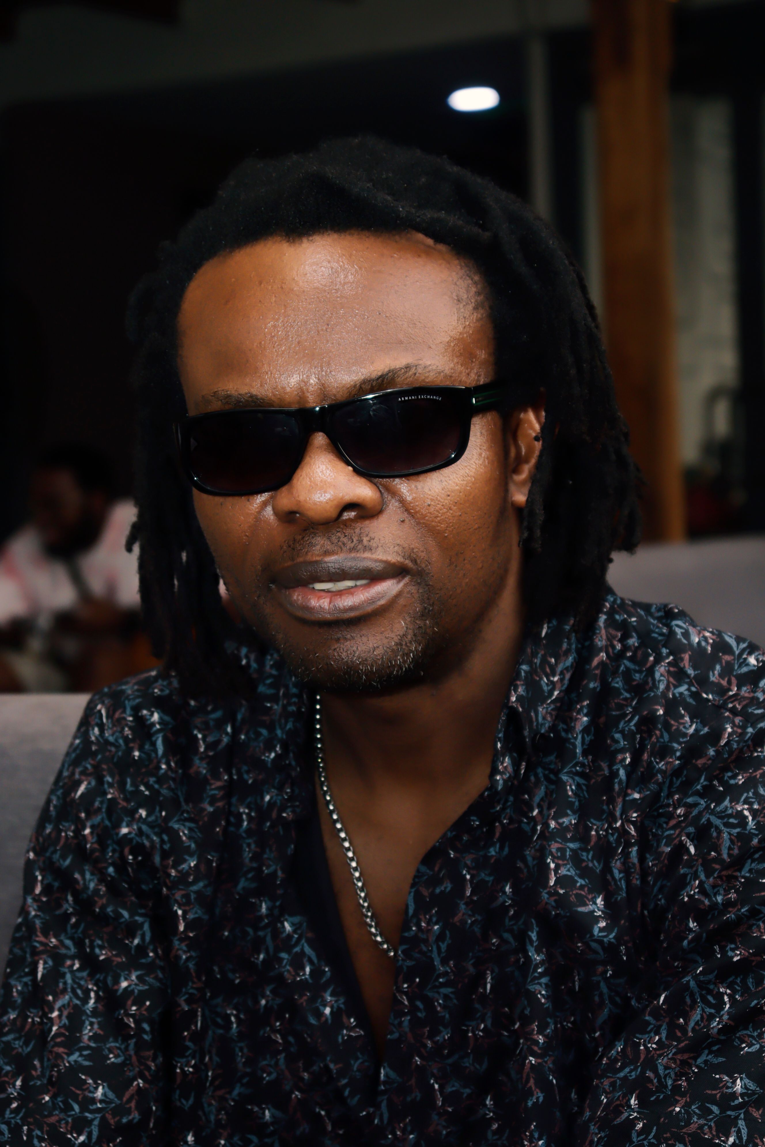 Stylish man sitting indoors, wearing sunglasses and a patterned shirt, exuding confidence and cool style.