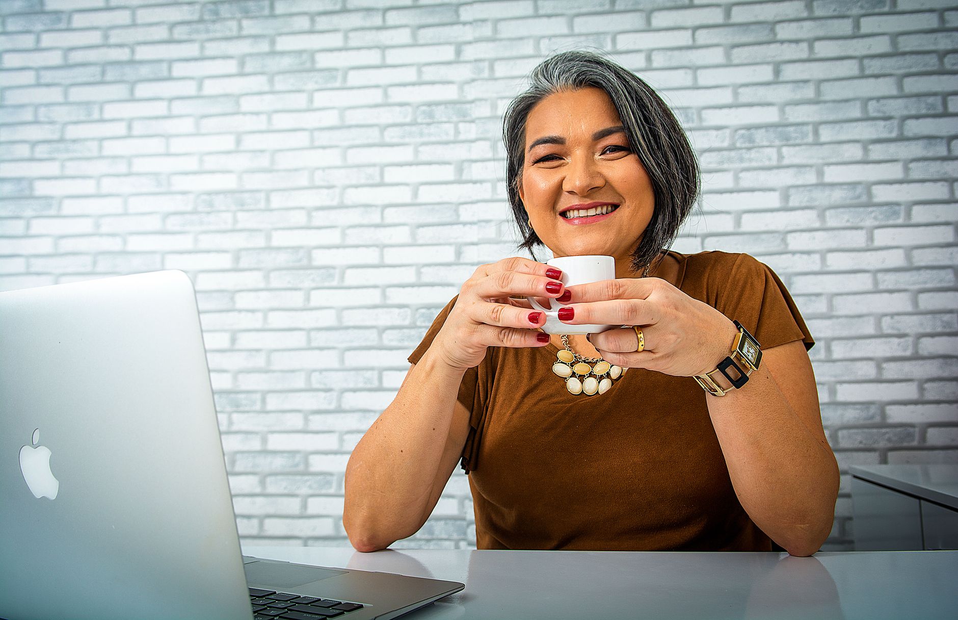 Middle-aged woman with gray hair enjoying coffee while working on a laptop, smiling warmly.