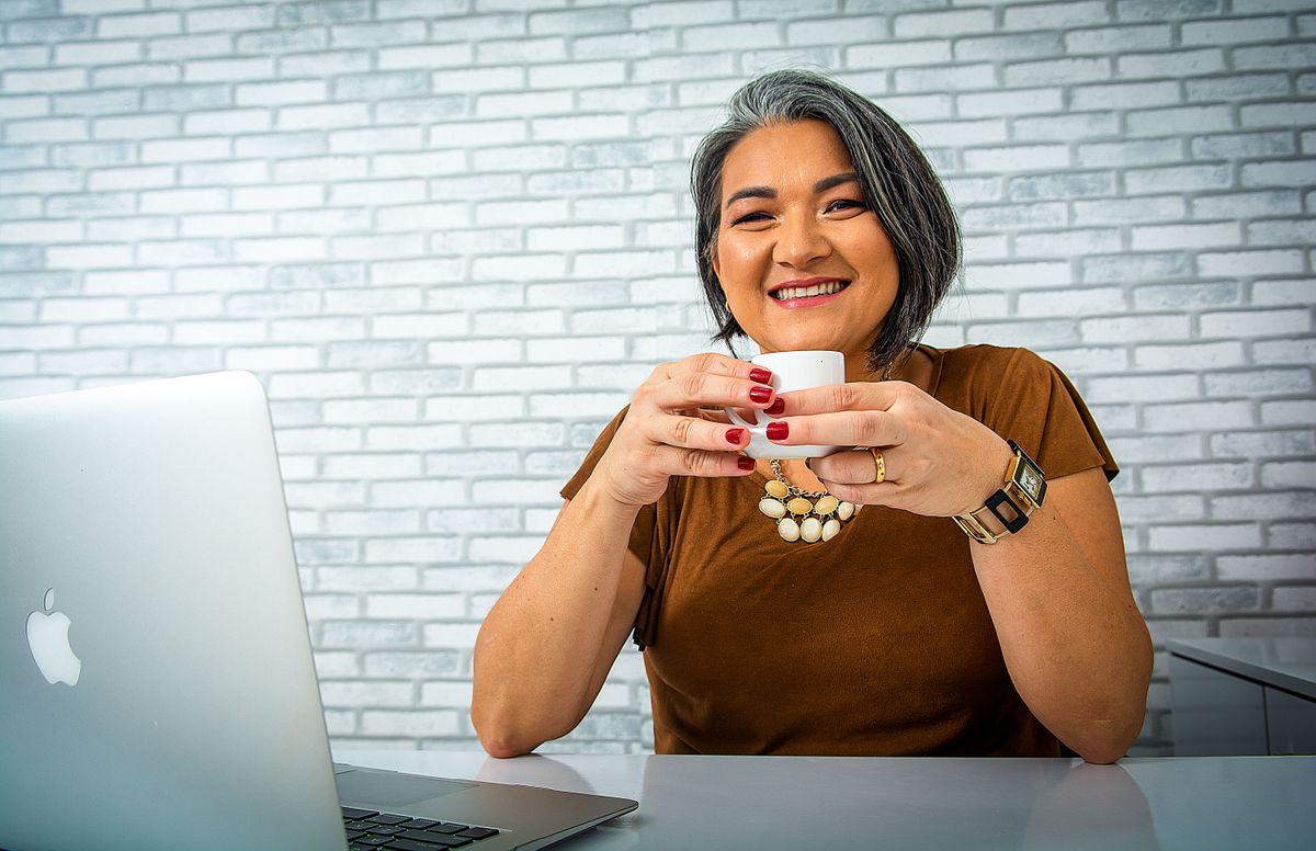 A coach smiling over coffee at her laptop at home