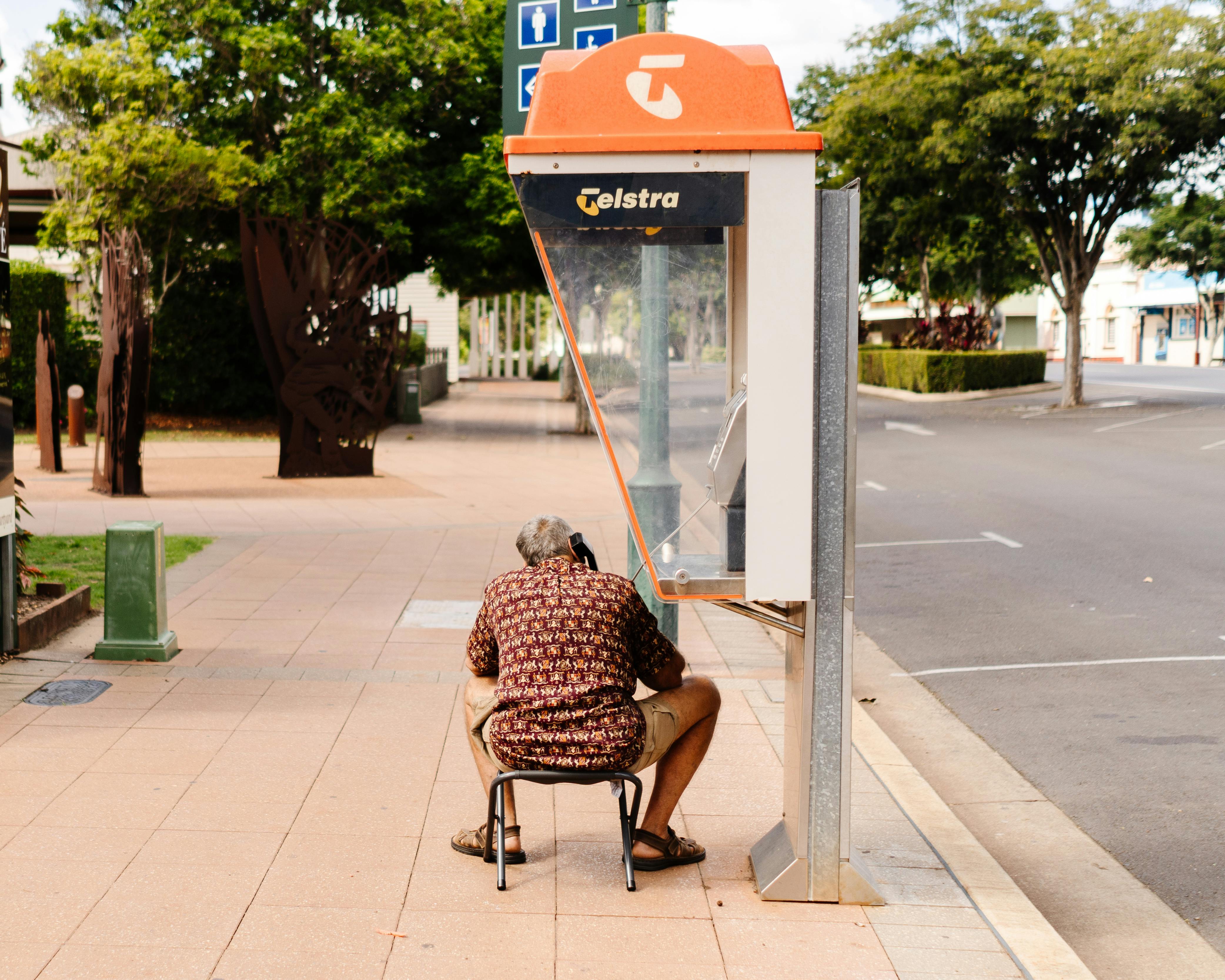 A man sits on a stool using a Telstra payphone in Childers, QLD, Australia.