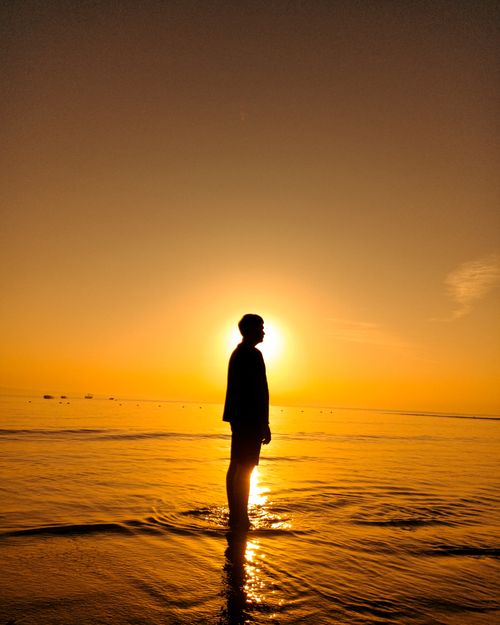 Free A man stands silhouetted against a vibrant sunset on a serene beach, reflecting over the sea. Stock Photo