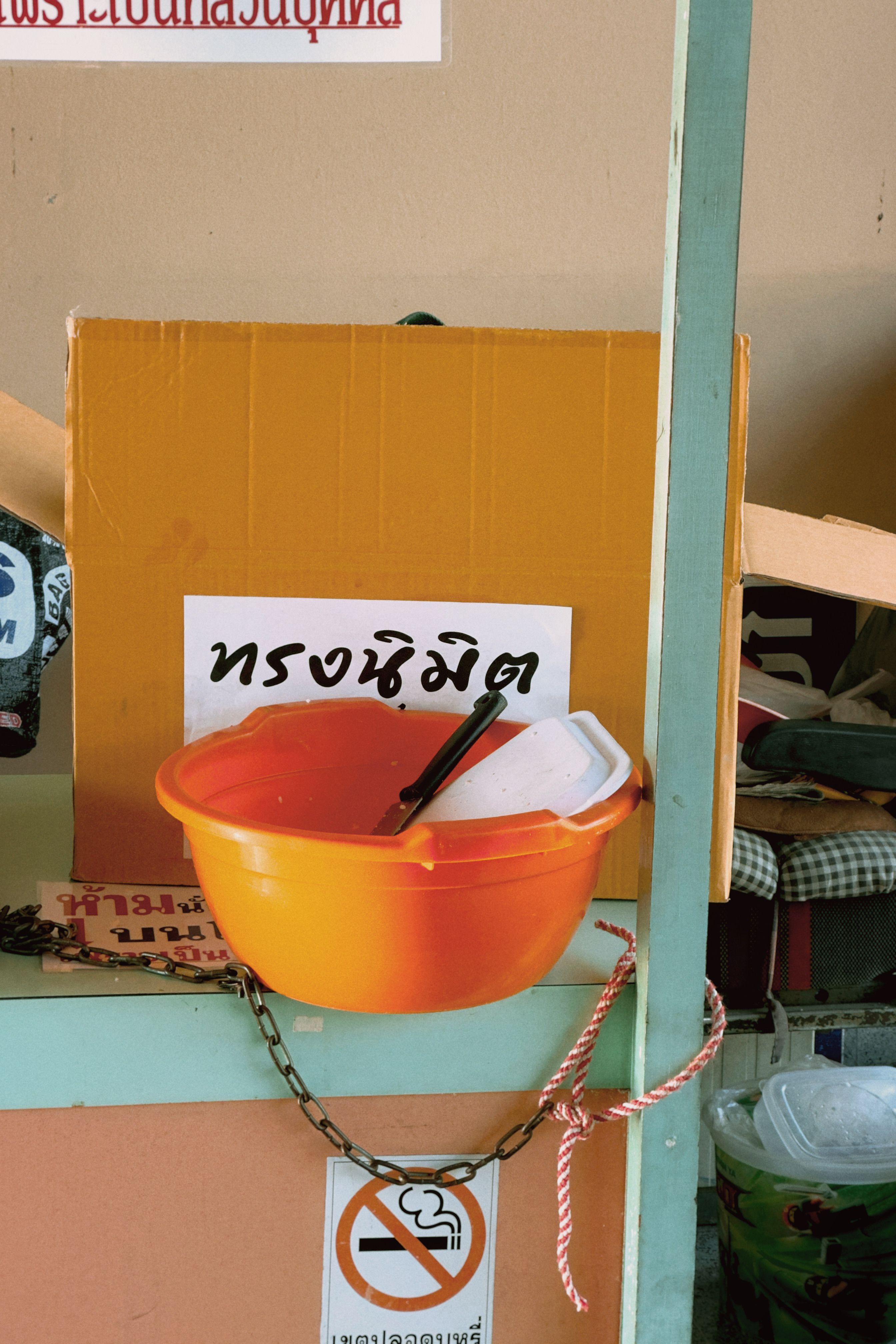 Bright orange bowl with utensils in a cluttered indoor storeroom with visible Thai language signage.