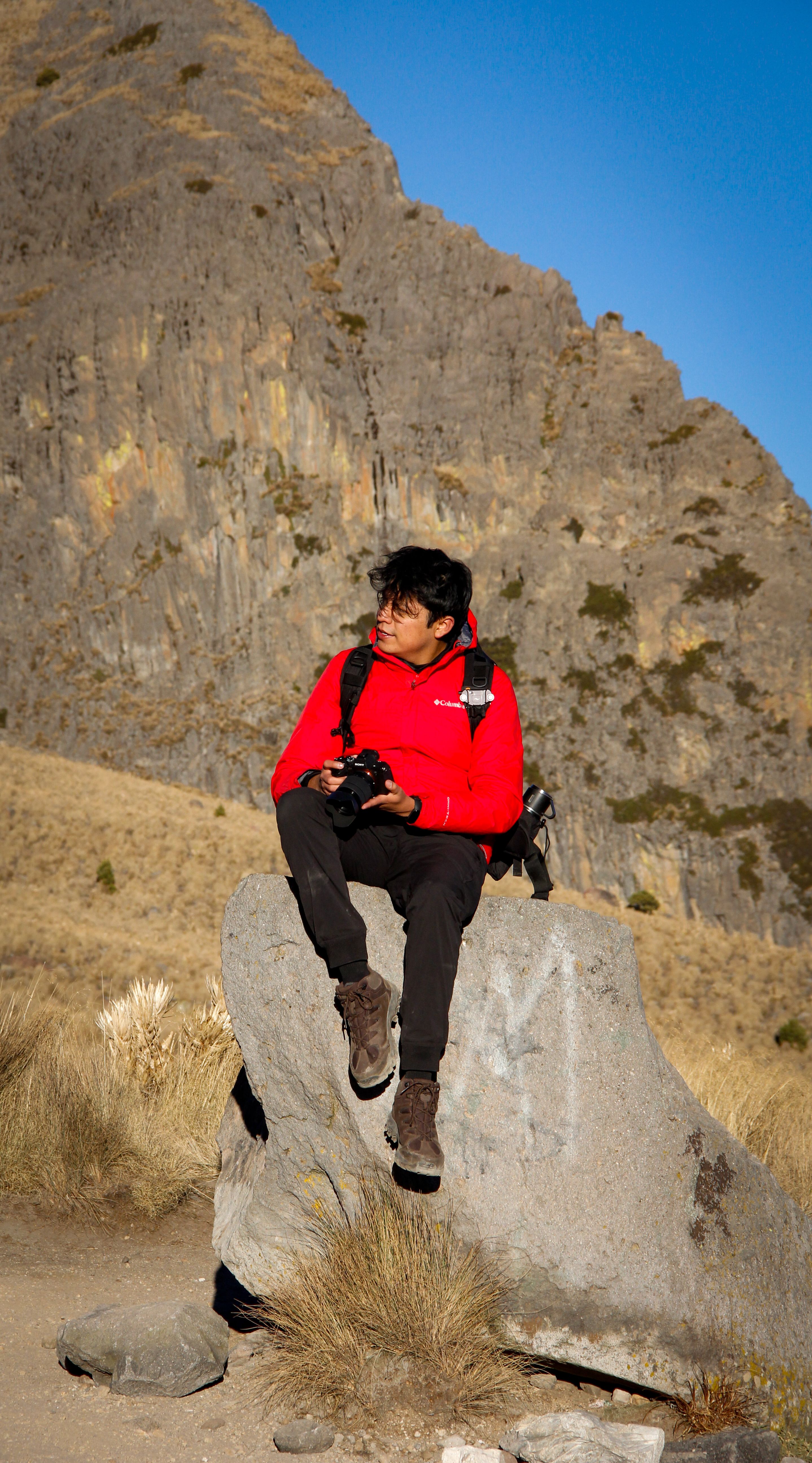 Young man in a red jacket sitting on a rock in the mountains of Mexico, holding a camera.