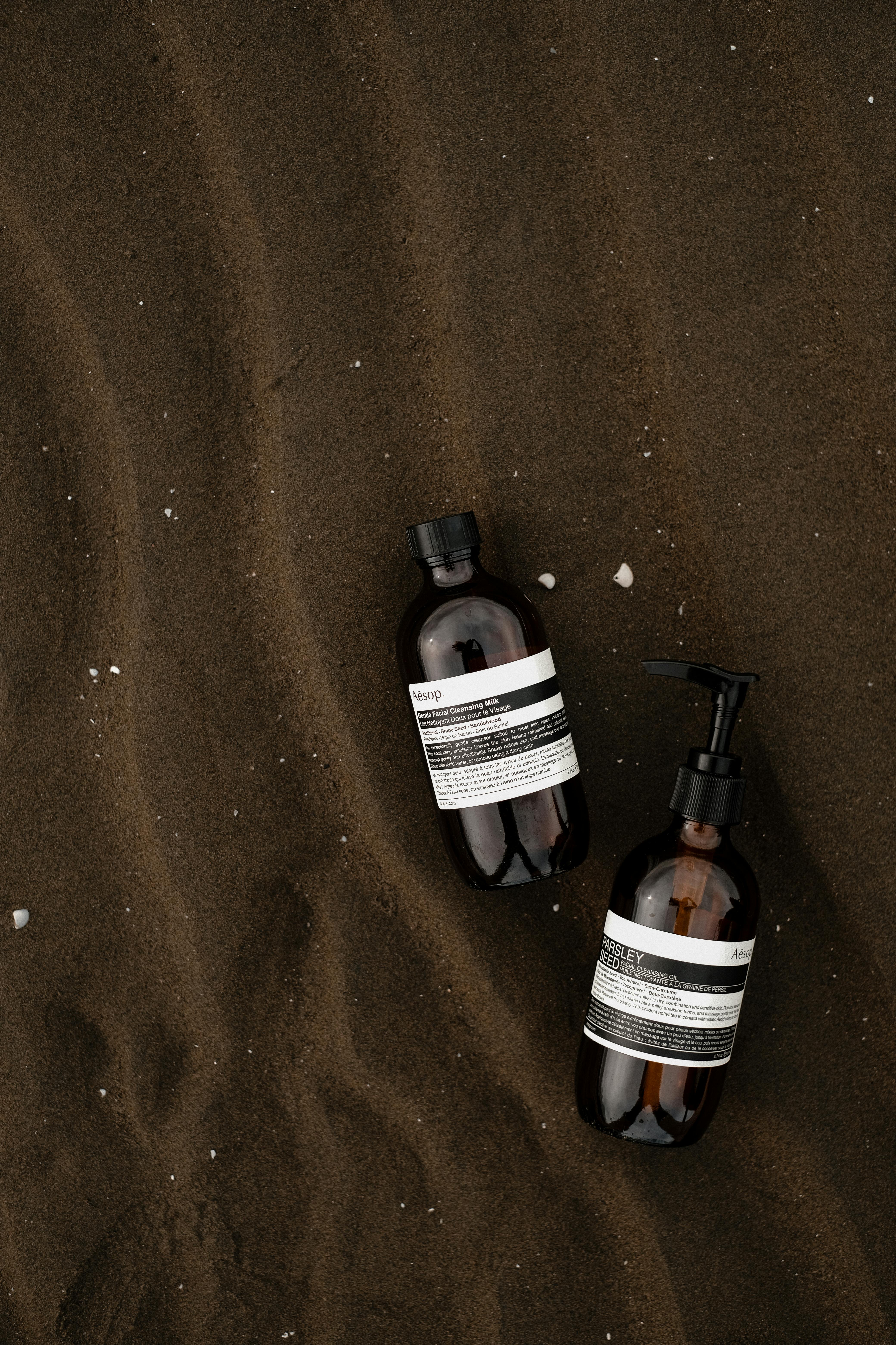 Two luxury cosmetic bottles on rippled dark sand, emphasizing elegance and simplicity.