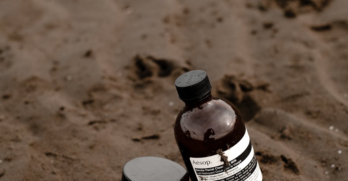 A bottle of sunscreen with a sun hat and sunglasses on a sandy beach.