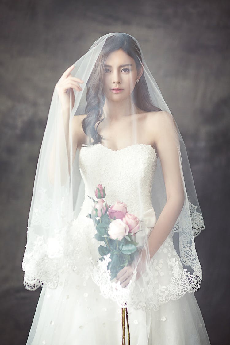 Woman In White Strapless Sweetheart Wedding Dress With Rose Bouquet Covered In White Veil