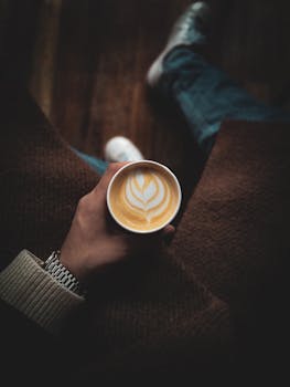 A hand holds a latte with beautiful foam art, captured from above in a warm, cozy setting.