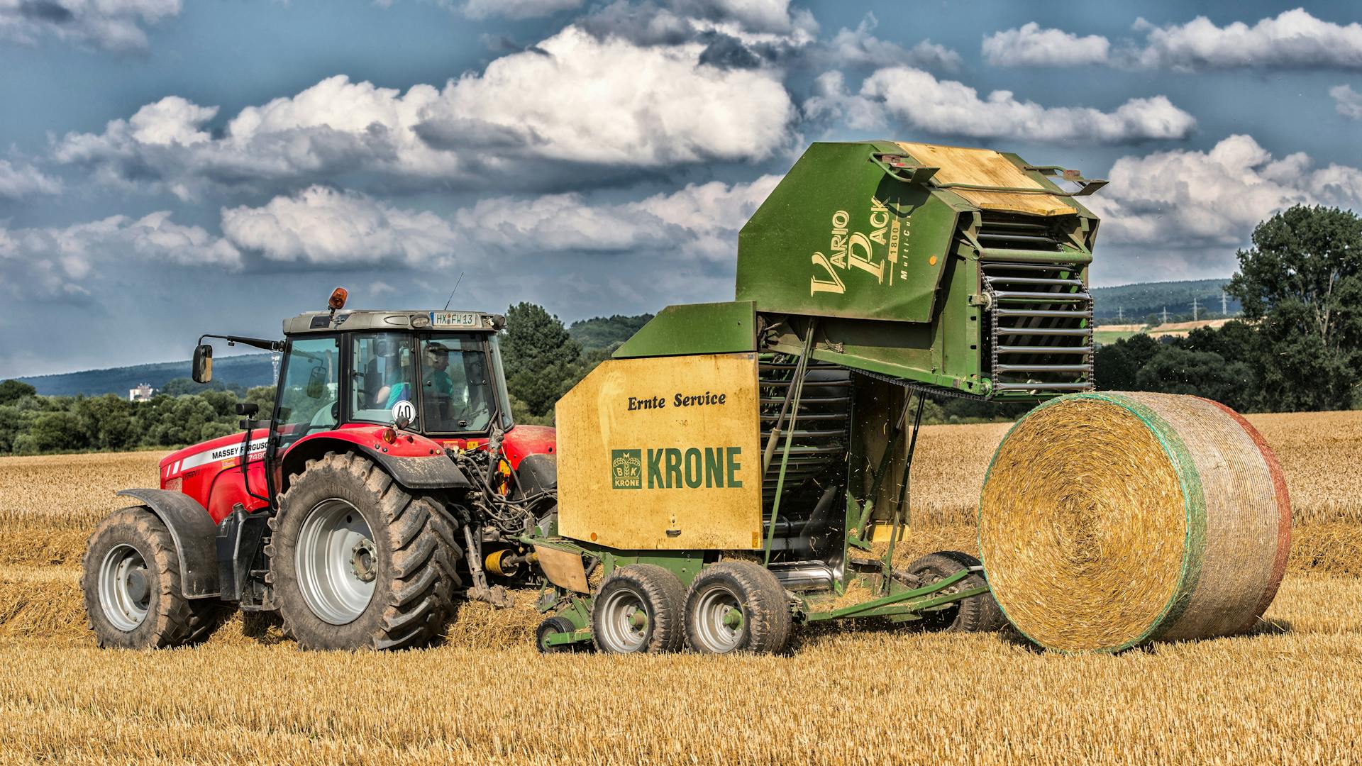 Rural scene of a red tractor and baler in a Höxter field, collecting hay under cloudy skies.