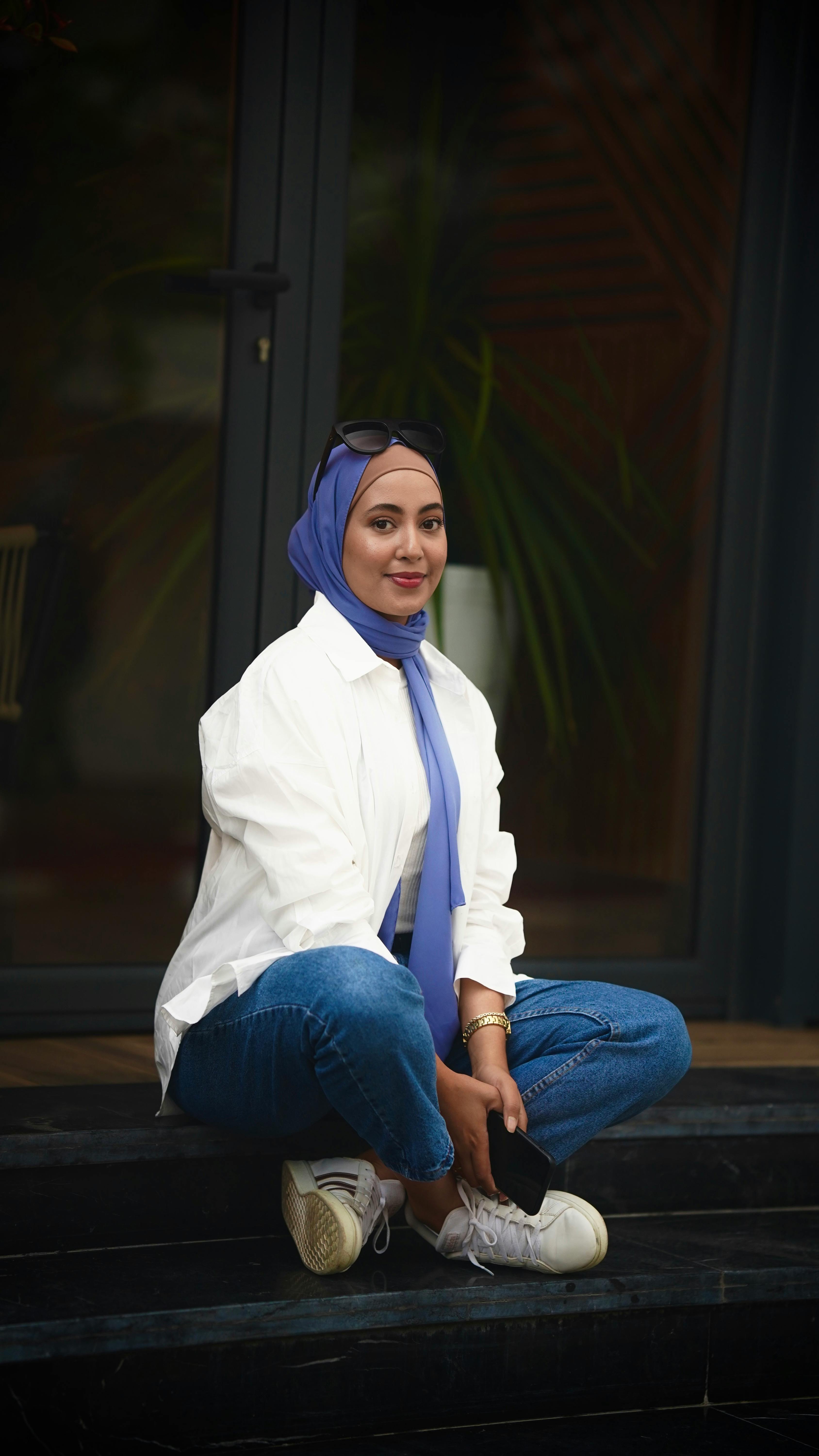 A fashionable woman wearing a blue hijab and casual attire, sitting on steps outdoors with a smile.