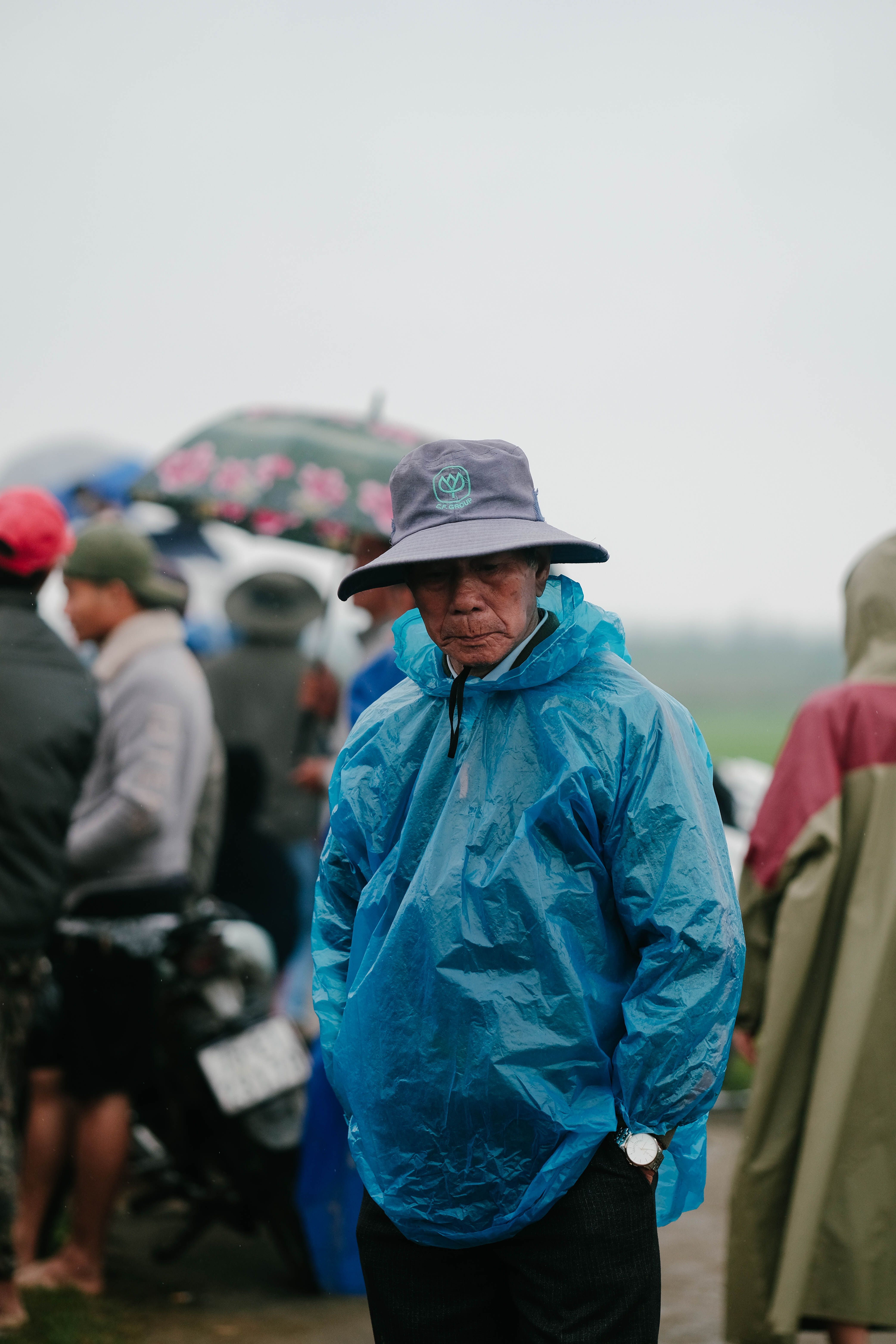 Portrait of an elderly man in a blue raincoat standing in a crowded outdoor setting during rain.