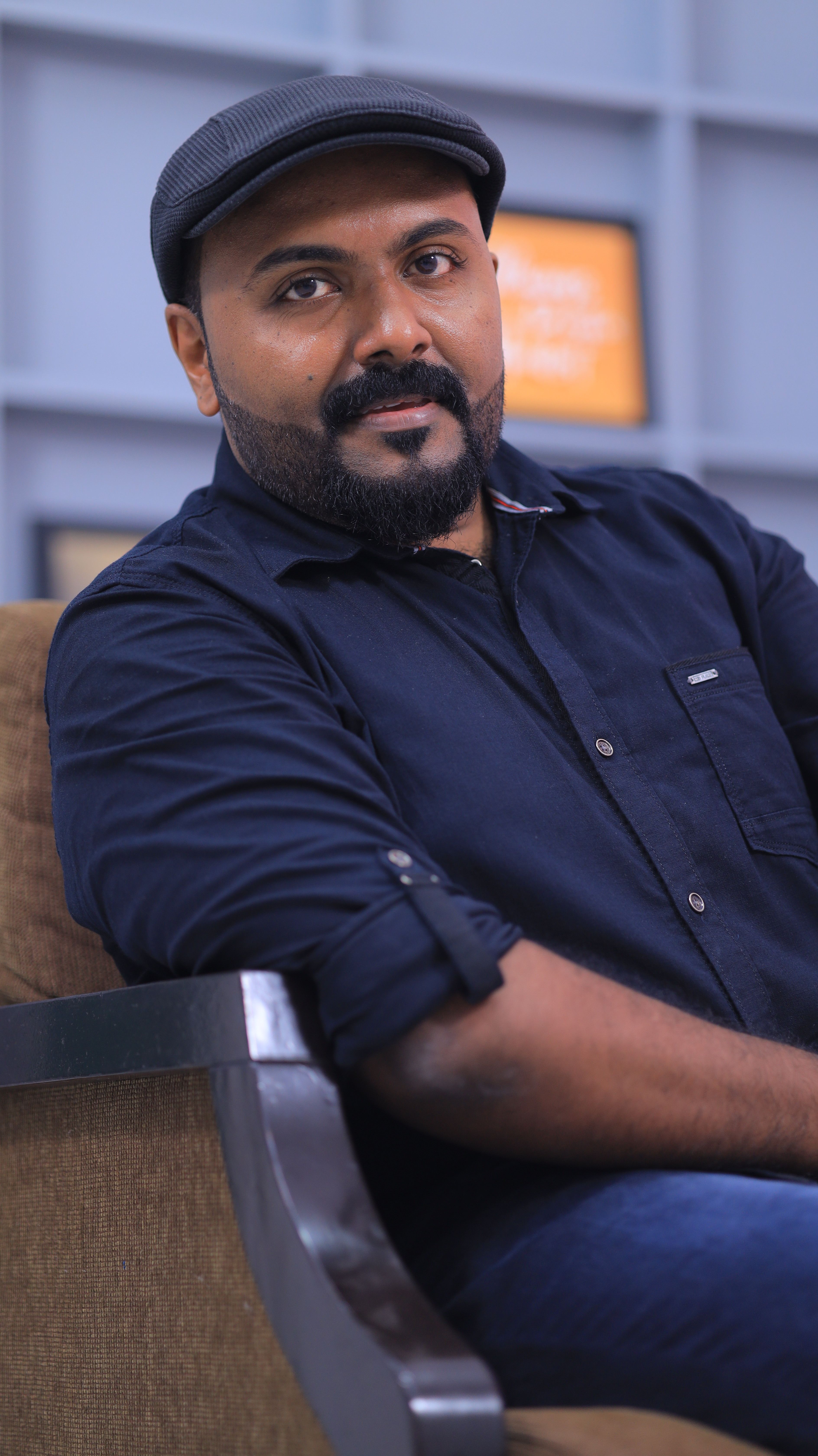 A relaxed portrait of a man sitting on a chair, wearing a navy blue shirt and a hat.