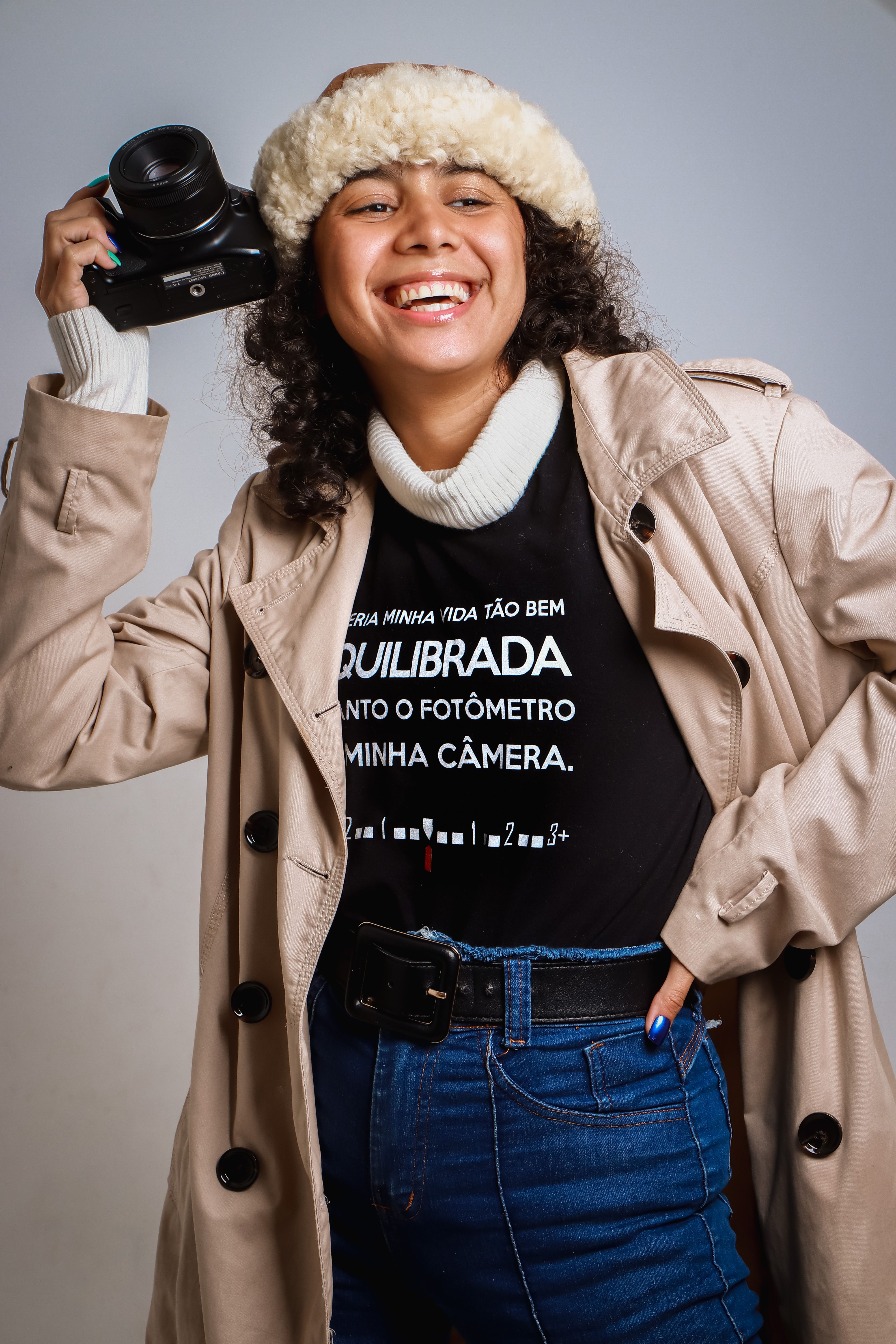 Smiling woman in trench coat holding a camera, embracing fashion and photography in SP, Brazil.